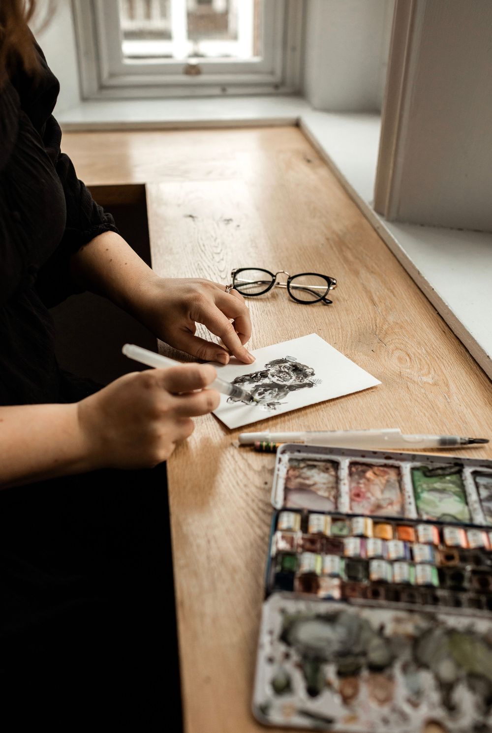 Artist sketching at wooden desk with watercolor palette and round glasses in natural window light.