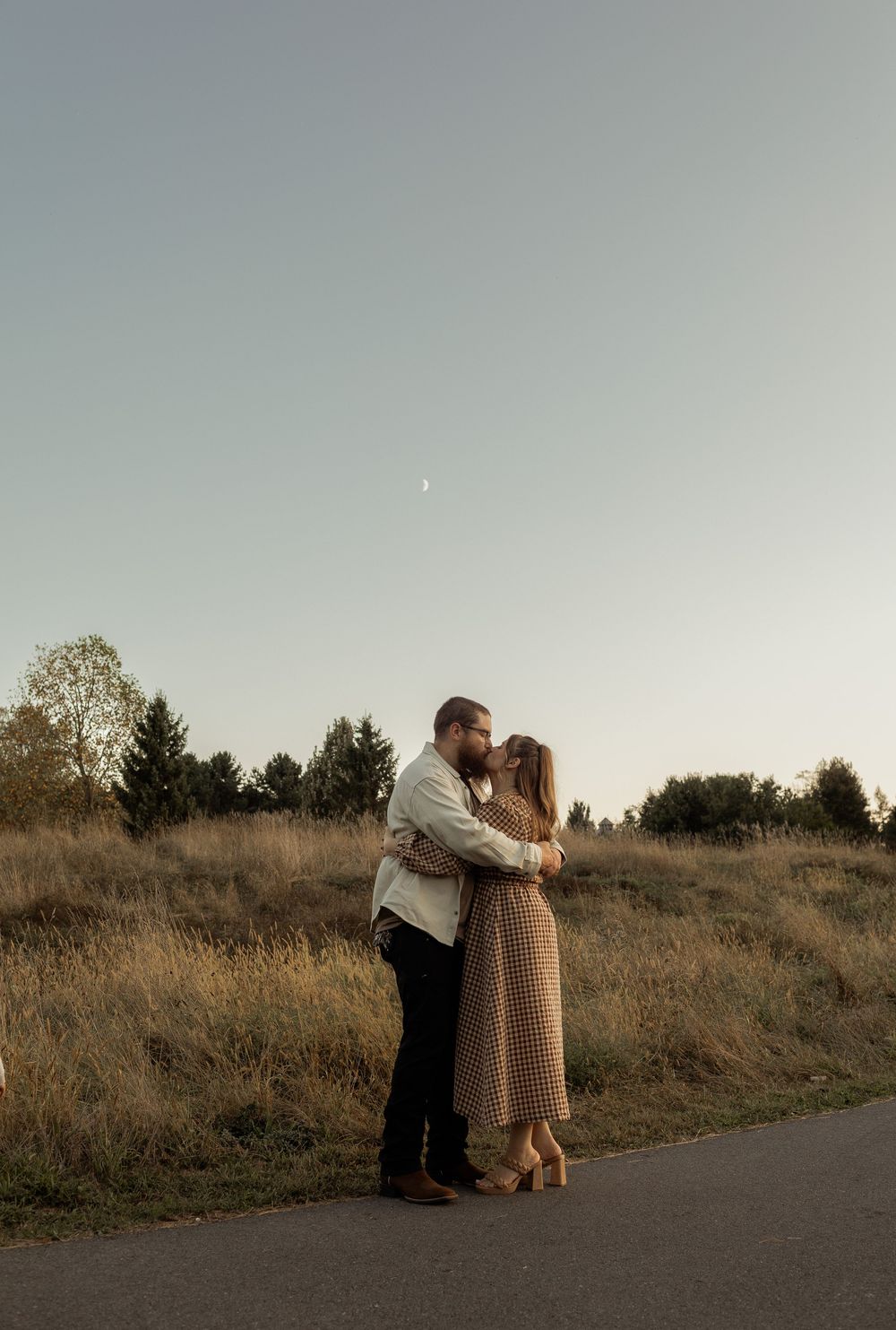 A romantic couple embraces on a path at dusk with evergreen trees and soft lighting in the background.