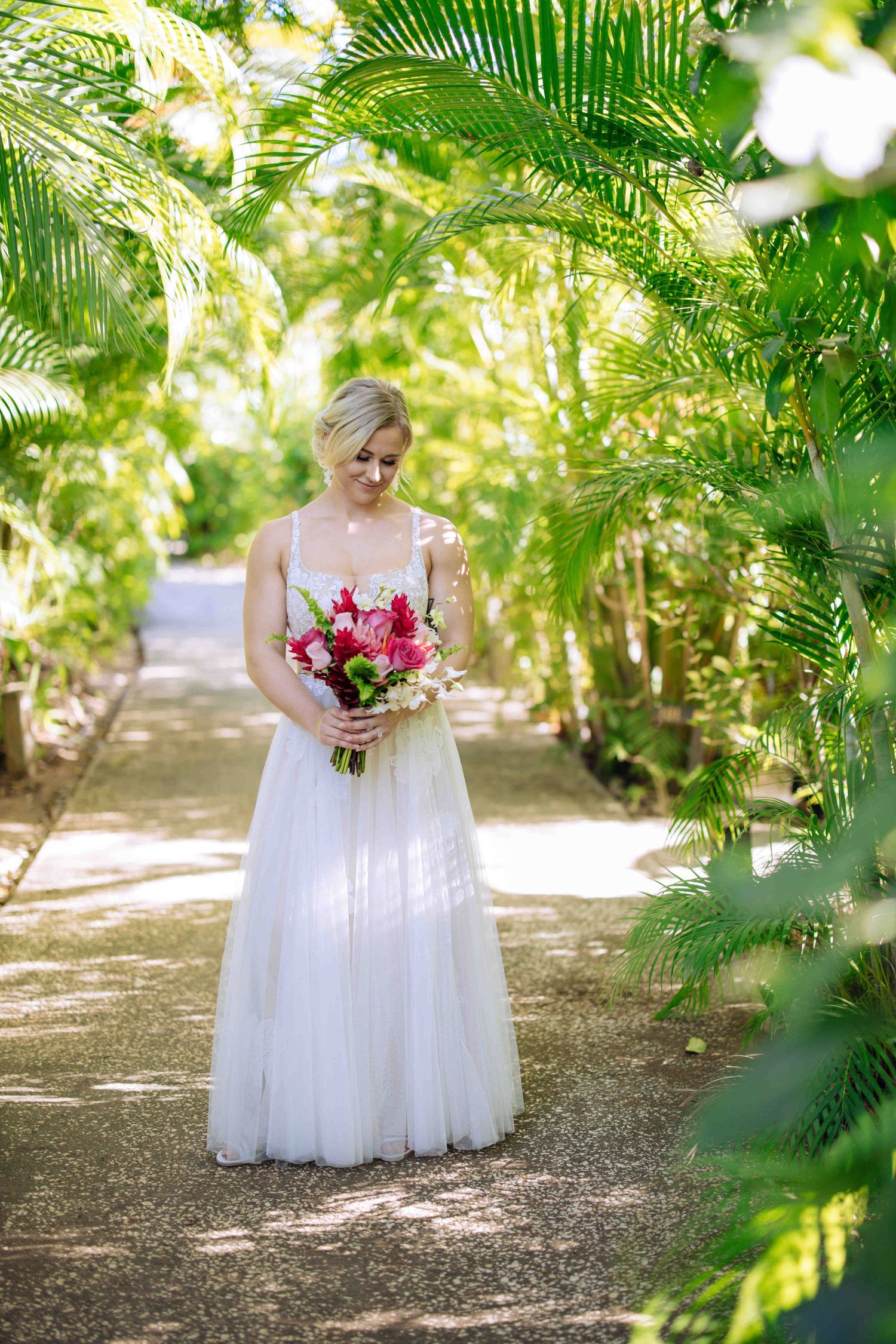 A bride in a flowing white gown holds a vibrant pink and red bouquet while standing among lush tropical palm trees.