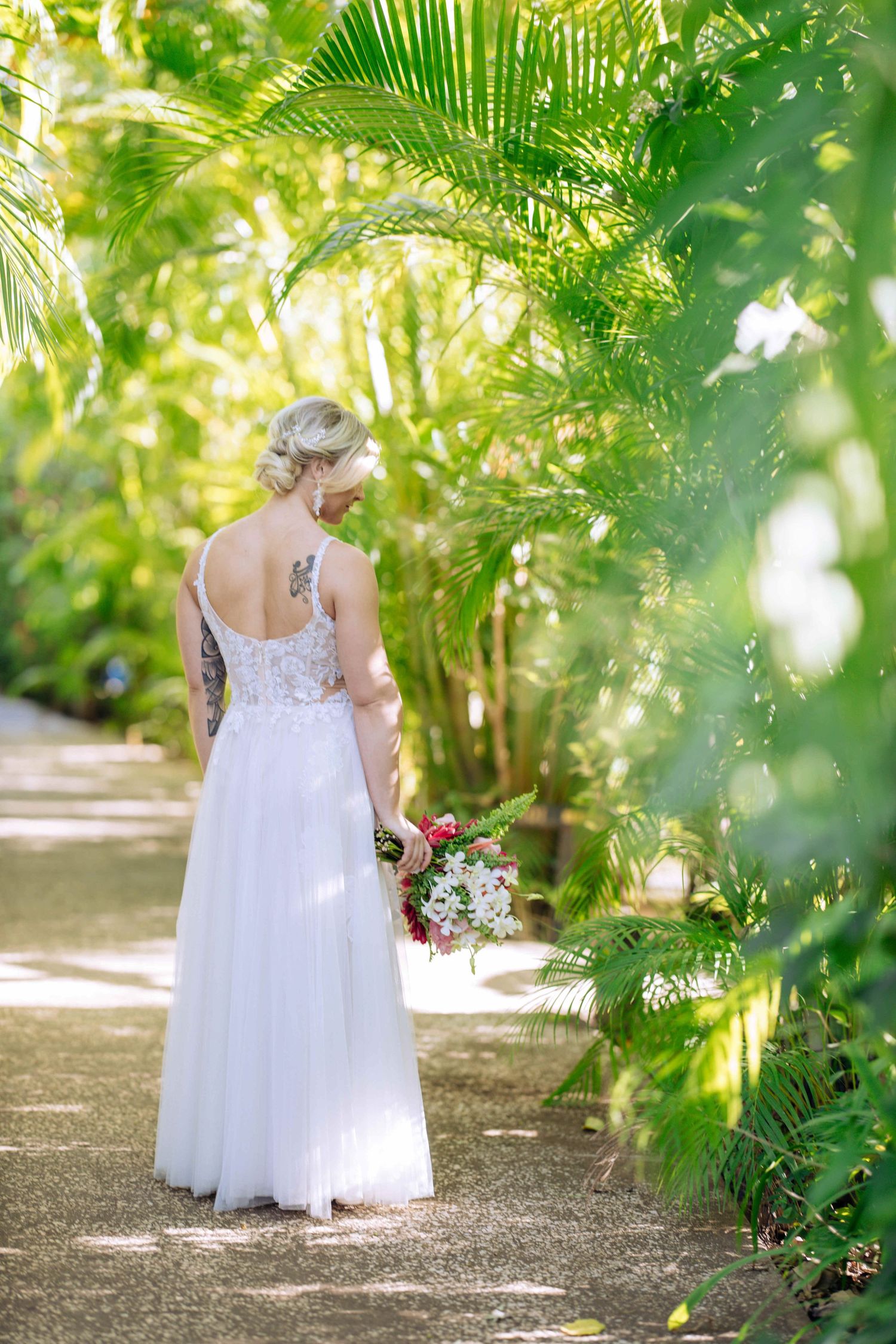 A white dress flows gracefully through a lush tropical garden path lined with palm fronds and greenery.