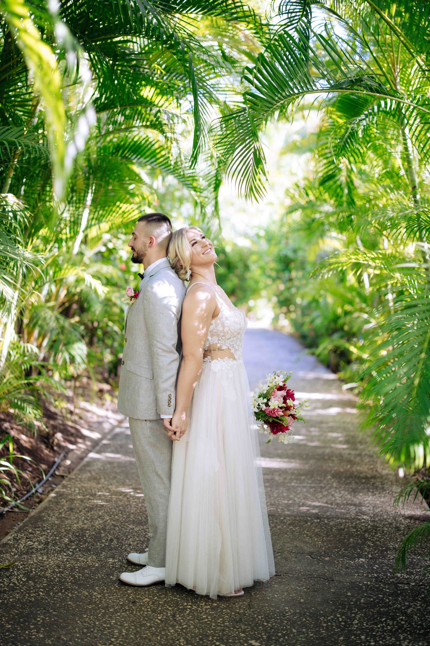 A couple in wedding attire stands together on a palm tree-lined path during their tropical destination wedding ceremony.