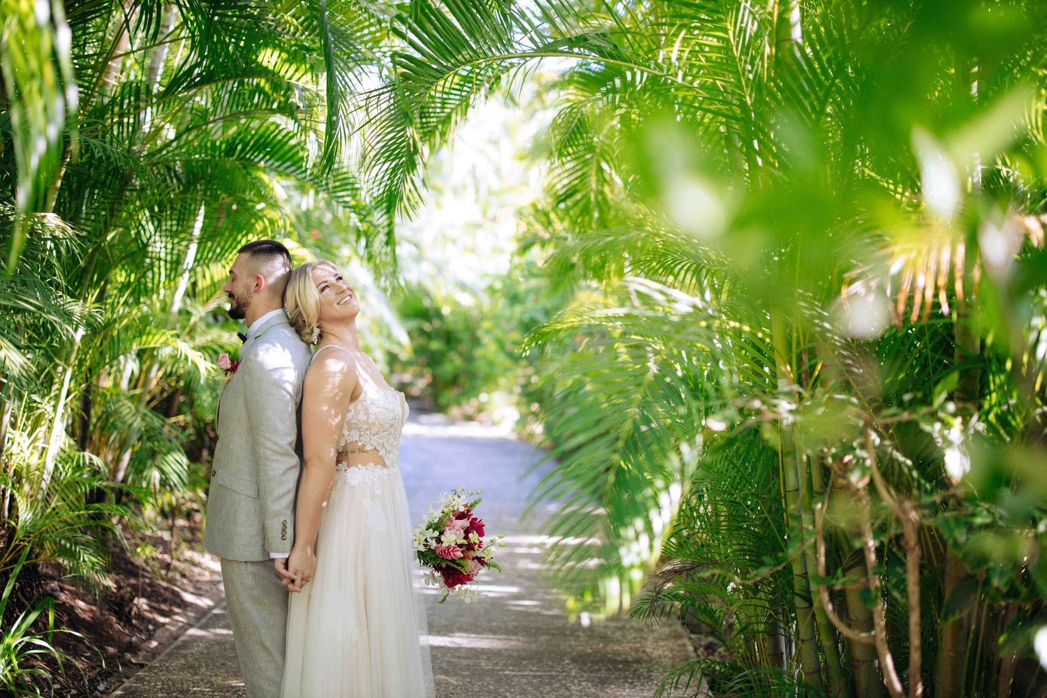 A couple embraces on a lush garden path surrounded by tropical palm trees and greenery on their wedding day.