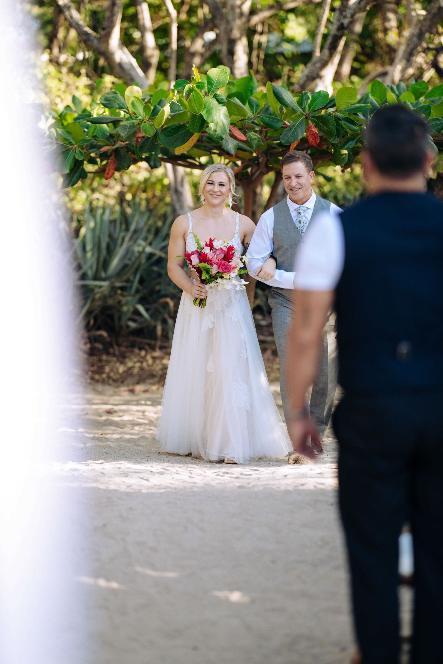 A beach wedding ceremony in progress with guests gathered under tropical foliage and sunlight filtering through trees.