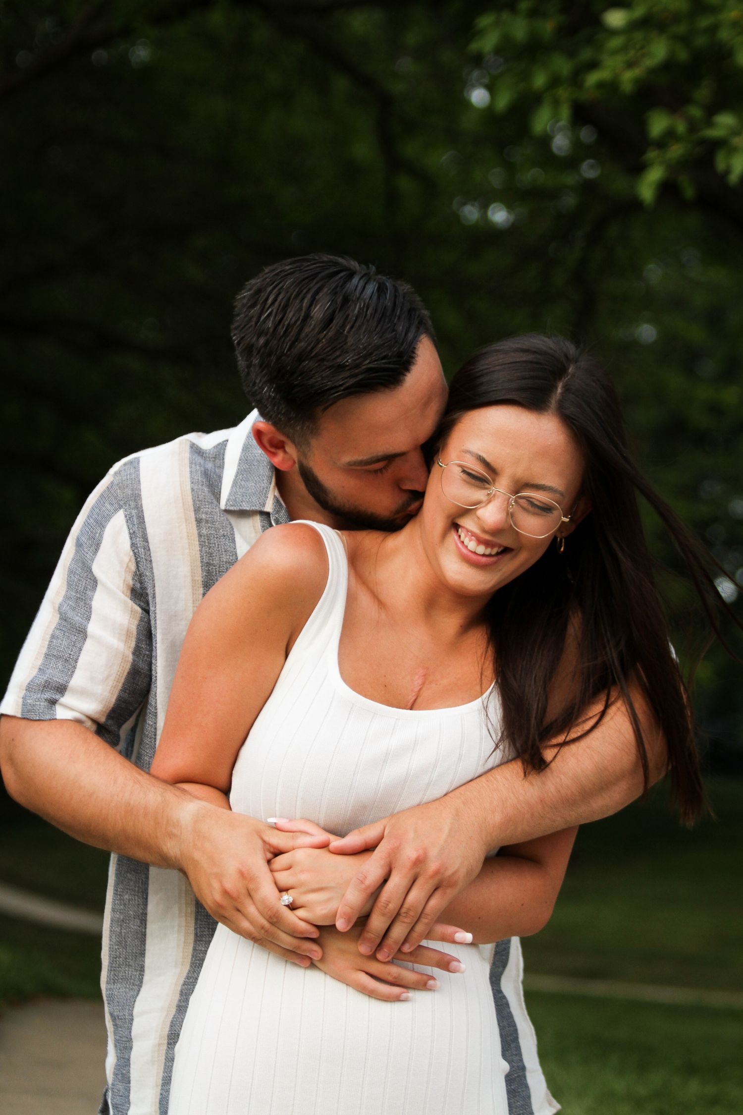 A woman scrunches up her face with laughter as her fiance hugs her from behind and kisses her on the neck in a playful image.