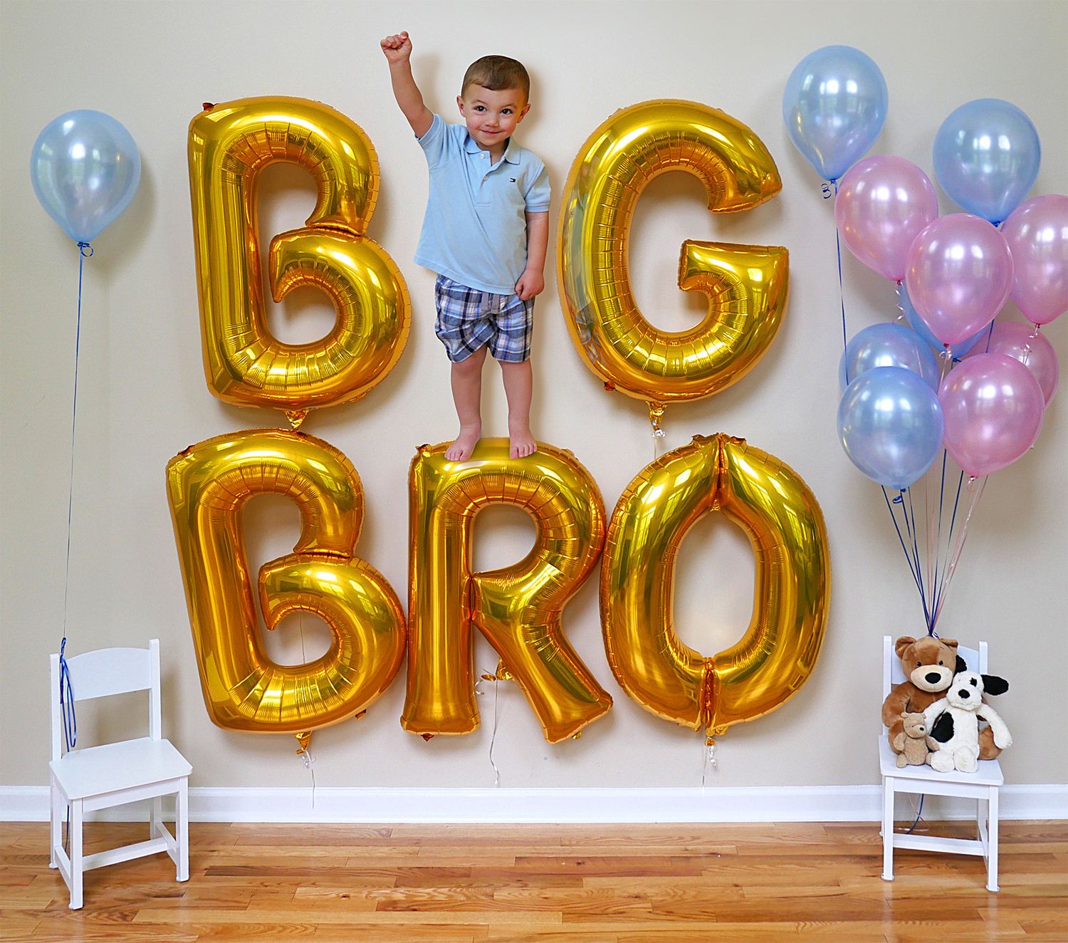 A birthday party decoration setup with gold letter balloons and blue and pink balloons against a white wall.