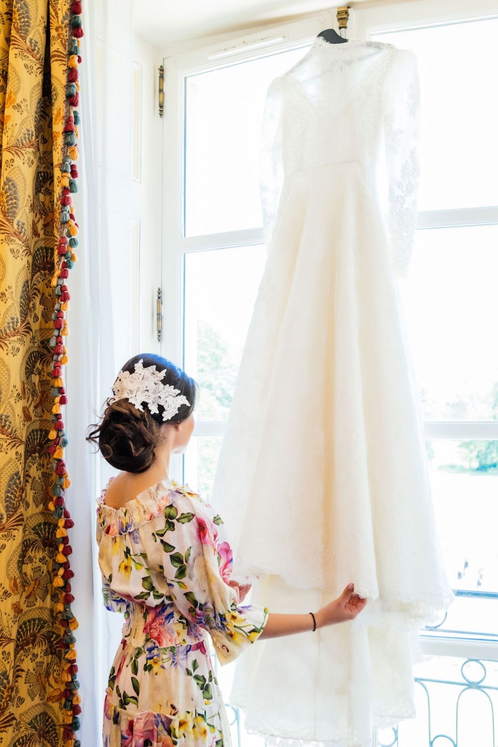Robe de mariée suspendue près d’une fenêtre, scène de préparation, détails délicats et lumineux.