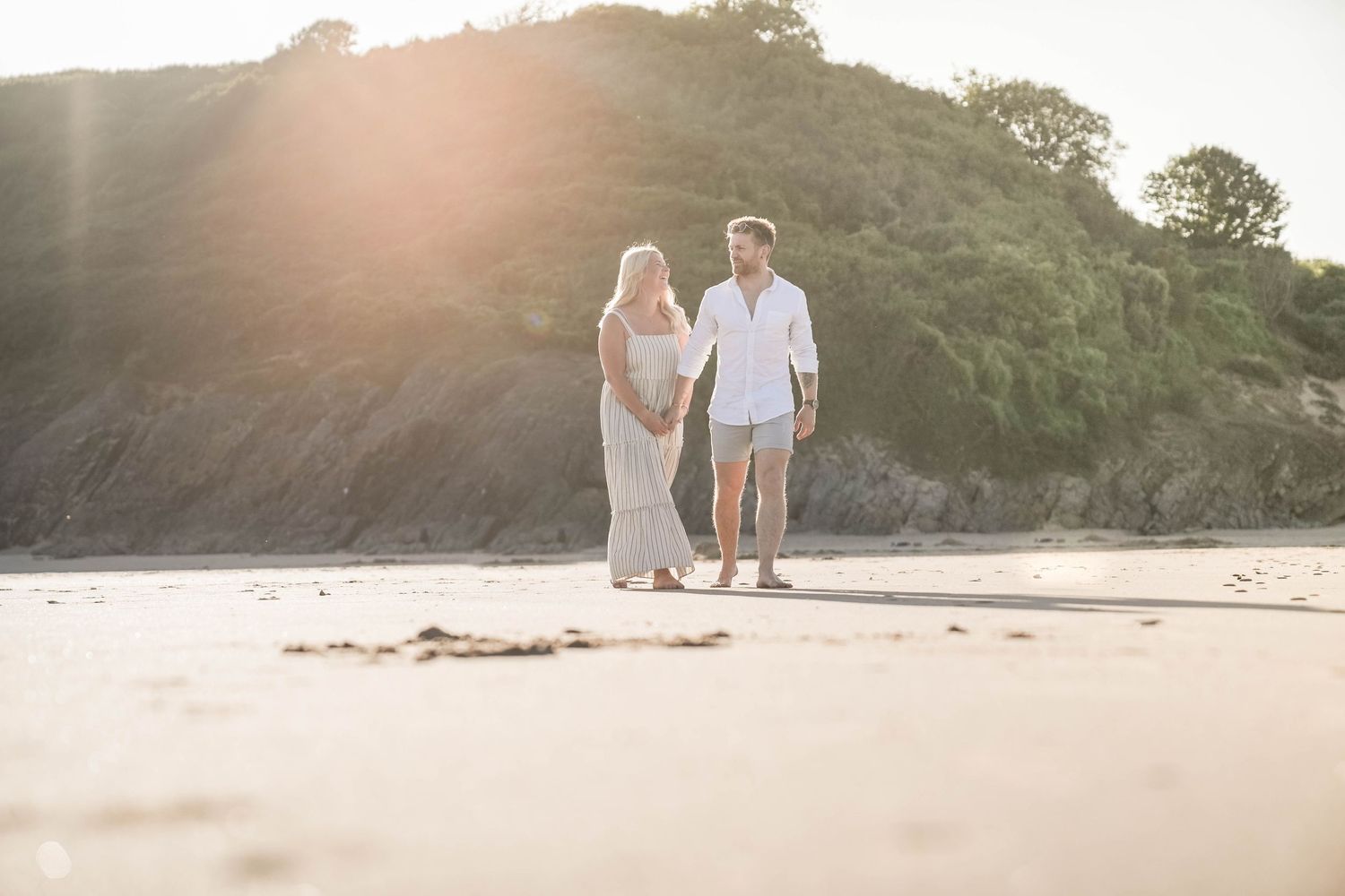 A couple walks hand in hand along a sunlit beach during golden hour as waves gently roll onto the shore.