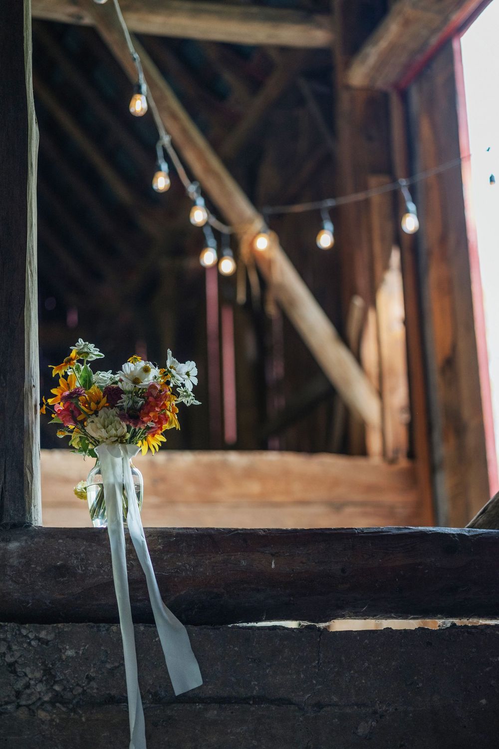 Small flower bouquets with white ribbons line a rustic wooden barn interior with string lights overhead designed by Pachamama.