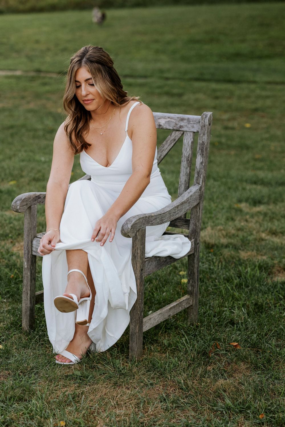 A bride in a white dress adjusts their shoes while sitting on a weathered wooden bench in a grassy meadow at Boyne Mountain.
