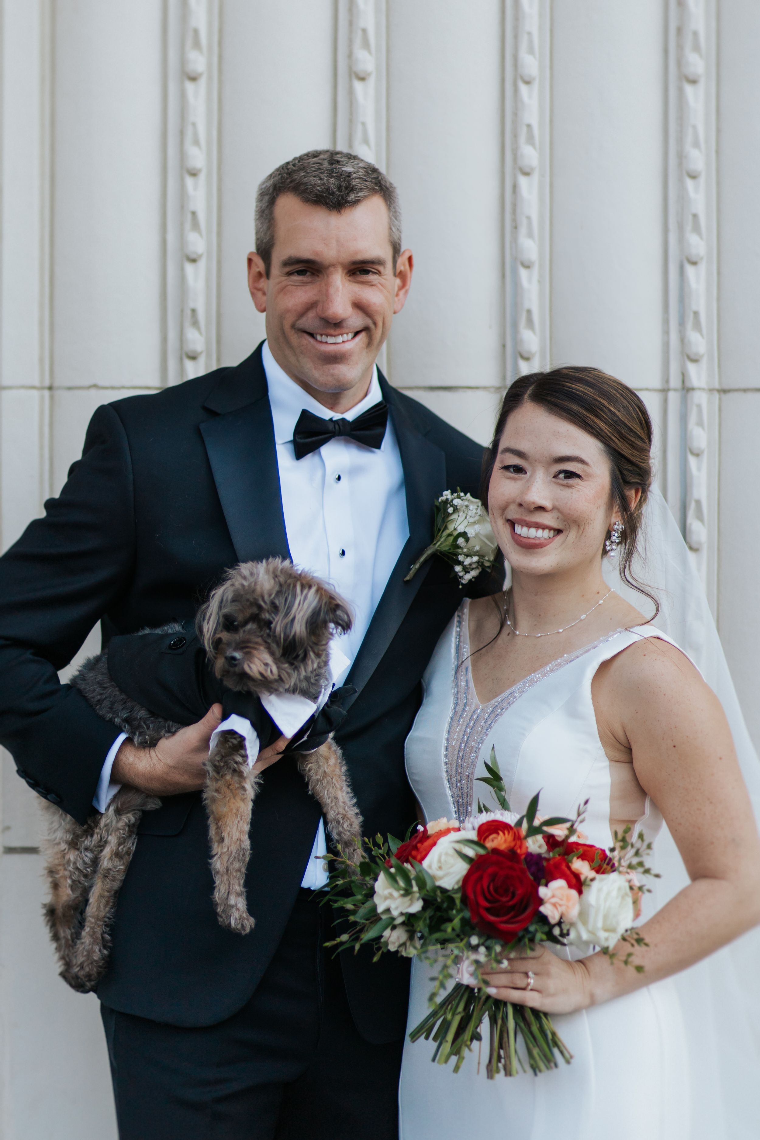 A happy couple in wedding attire poses with their small brown dog against a white wall background.