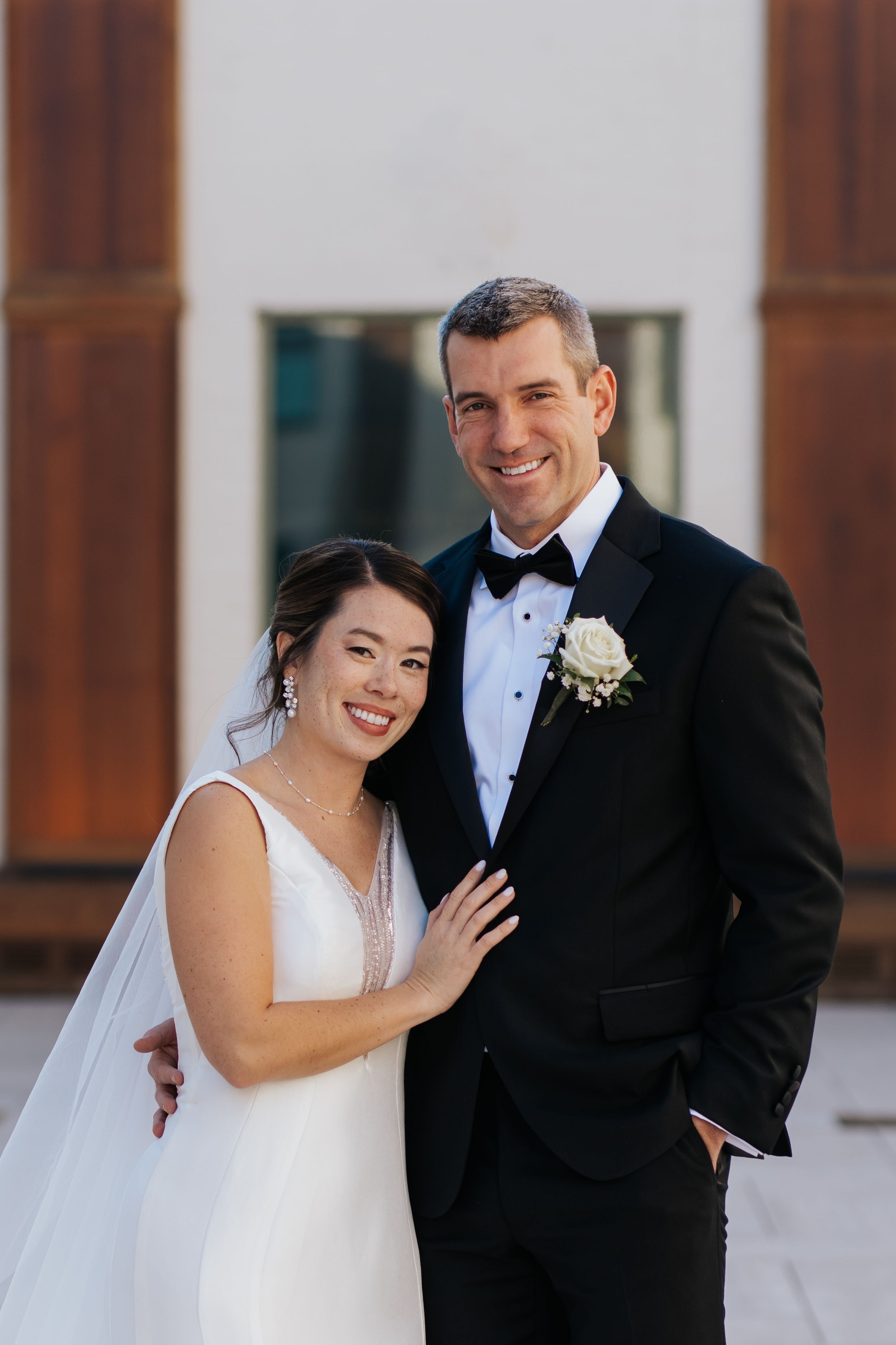 Happy couple shares joyful moments in formal wedding attire against a light colored wall.