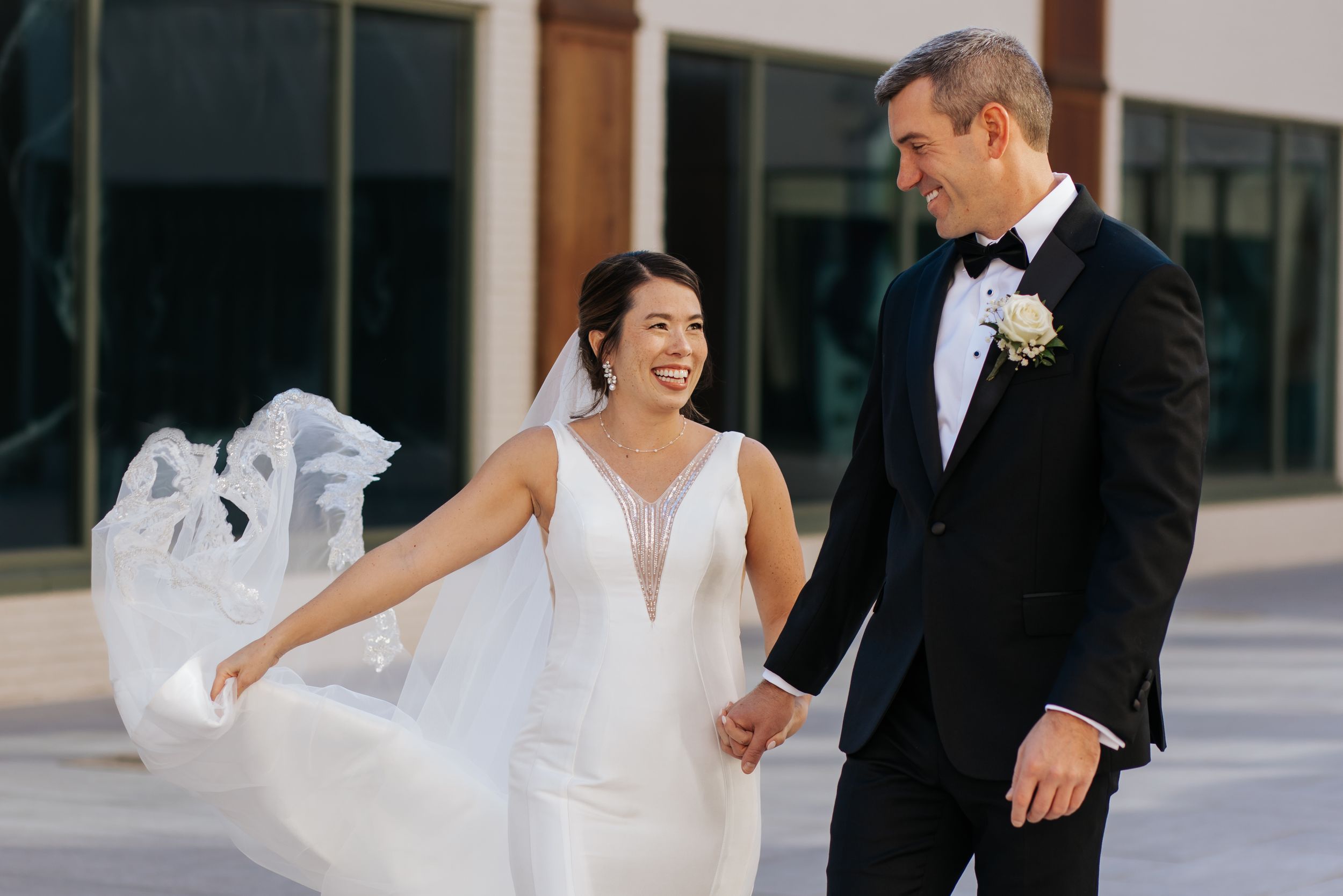 A joyful wedding couple laughs together outdoors, with the bride's veil flowing in the breeze.