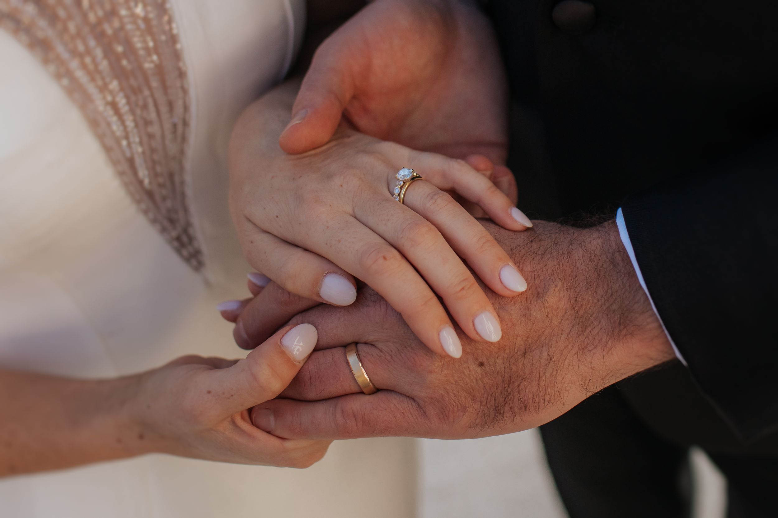 Close-up shot of newlyweds' hands with wedding rings intertwined against dark fabric background.