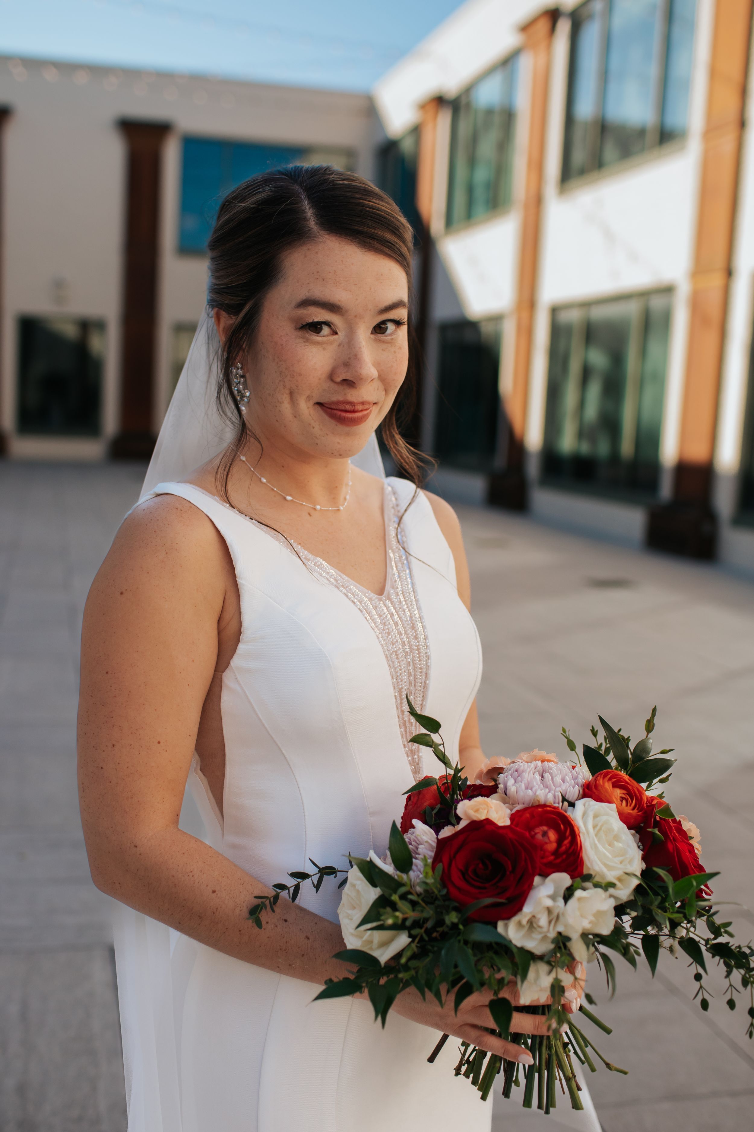 A bride in a sleeveless white dress holds a red and white rose bouquet during her wedding day.