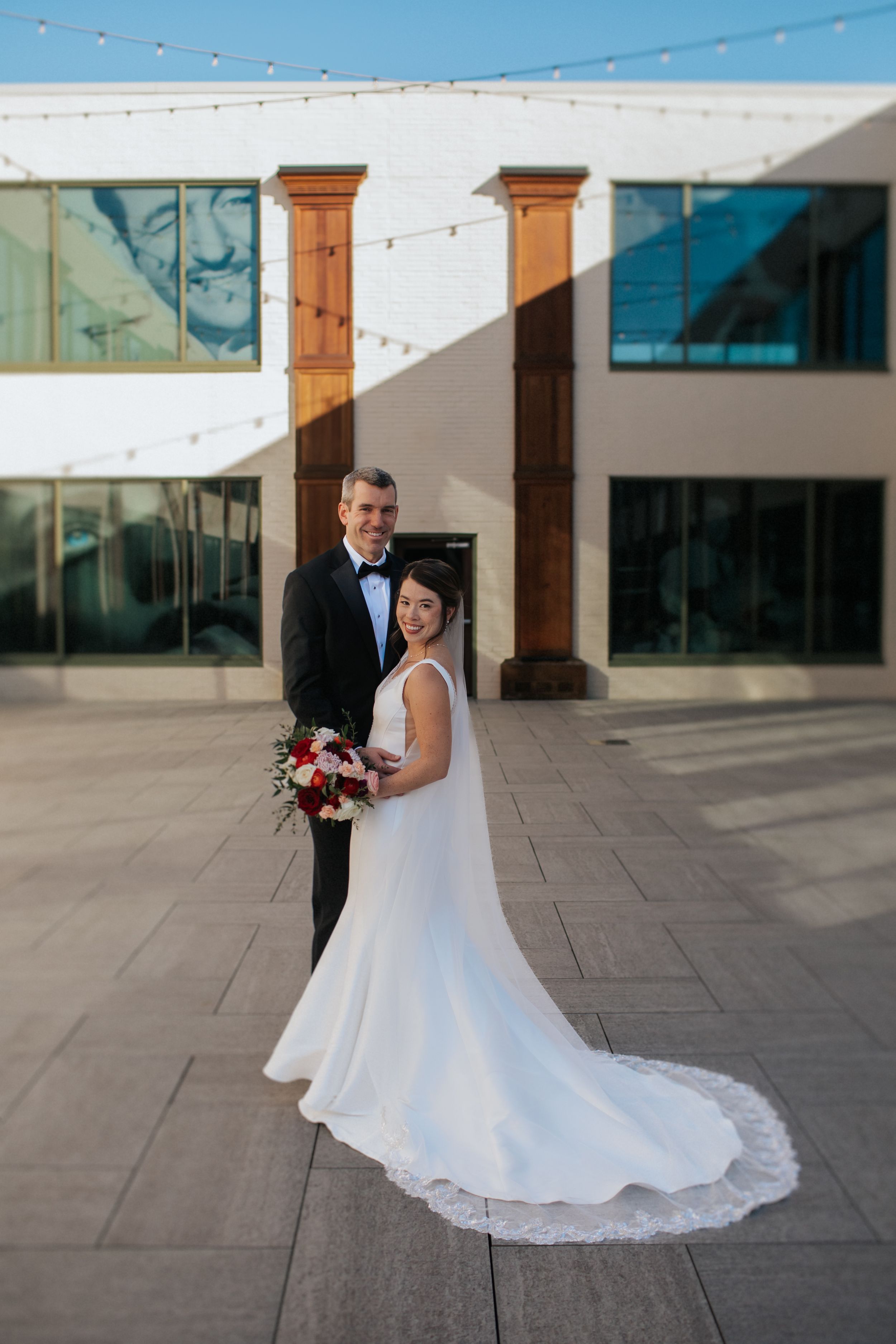 A wedding couple poses on a modern patio with string lights and architectural columns in the background.