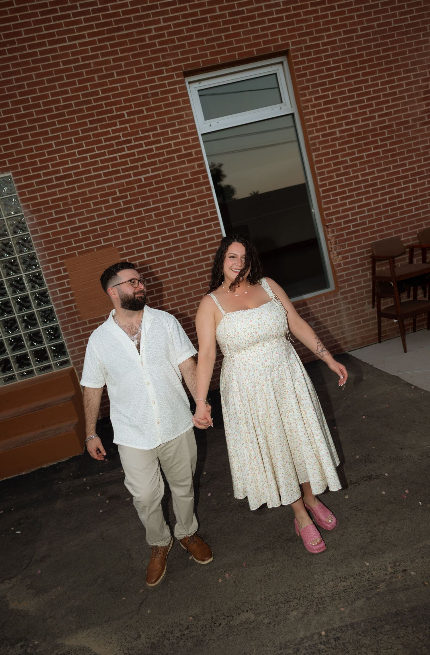 A couple in white and cream outfits hold hands while walking together in front of a brick building.