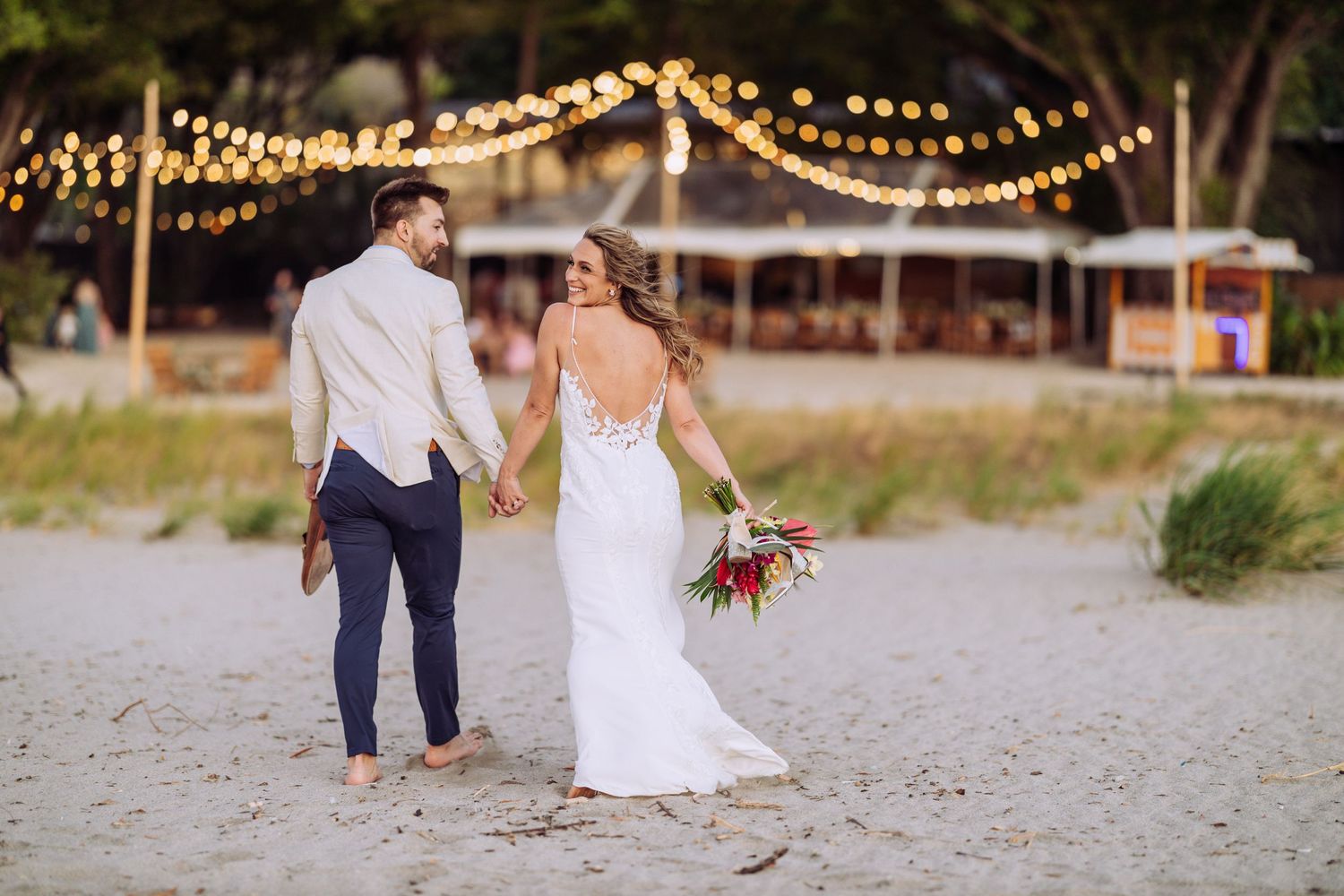 Newlyweds walking on beach at sunset under string lights.