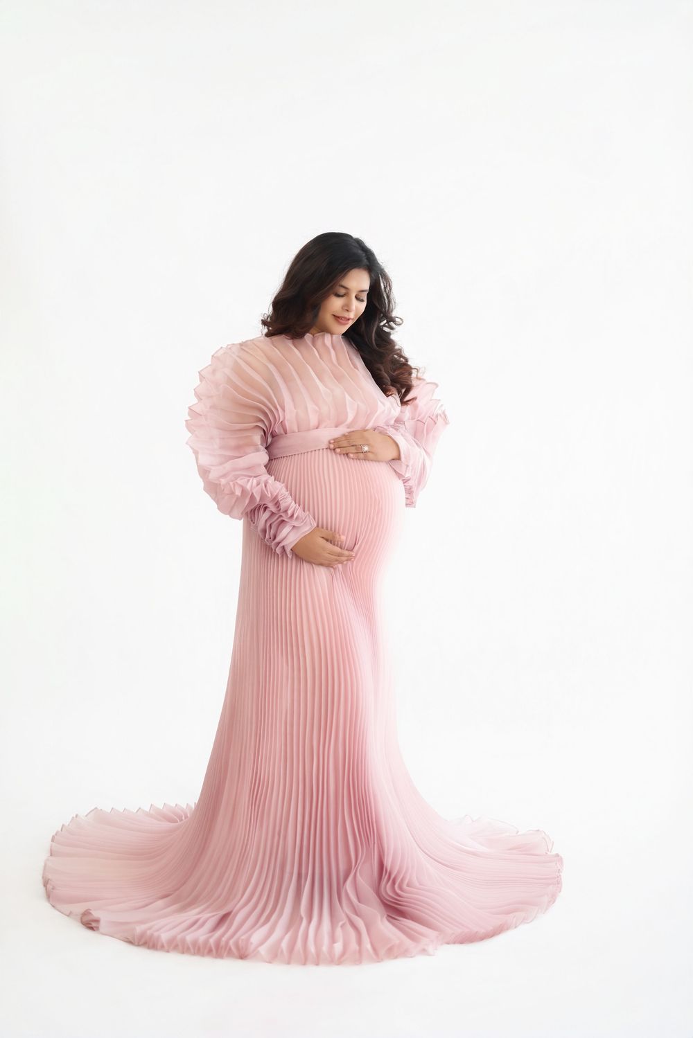 Woman wearing flowing pink pleated maternity gown poses gracefully against white backdrop in studio setting.