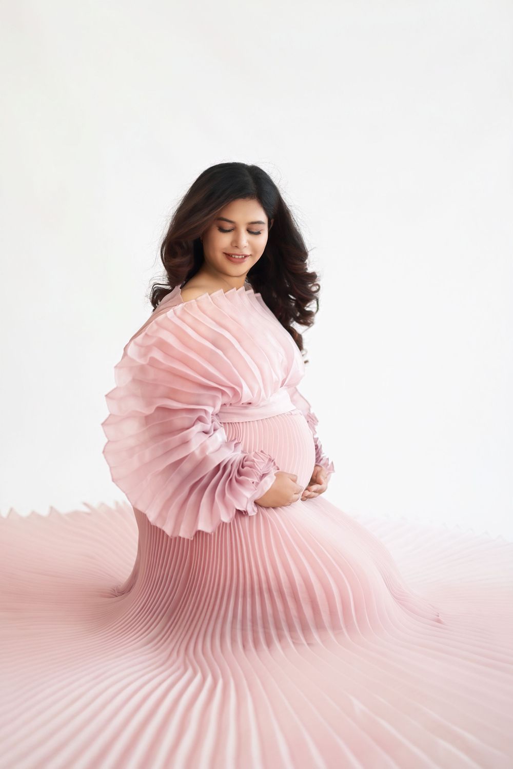 A woman in a flowing pink pleated dress poses gracefully against a white backdrop in a creative fashion photo series.