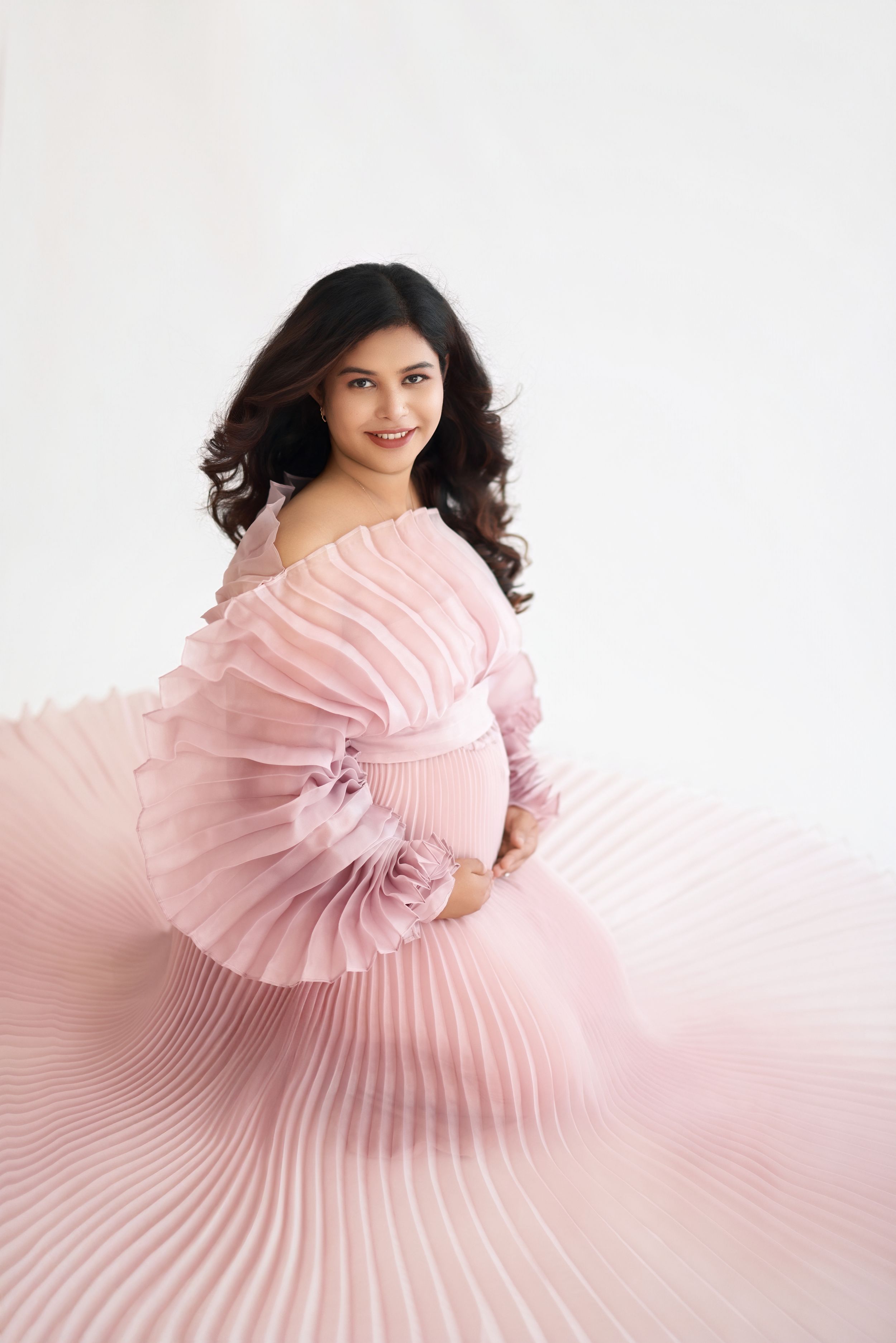 Elegant maternity photo featuring a flowing pink pleated gown against a white backdrop.