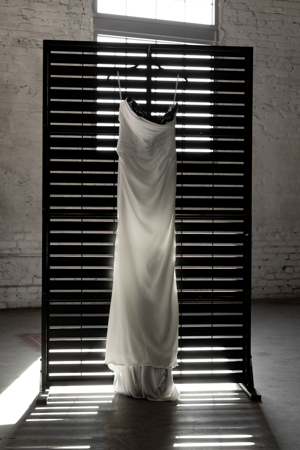 Wedding dress hanging in front of slatted window blinds creating dramatic lighting and shadows.