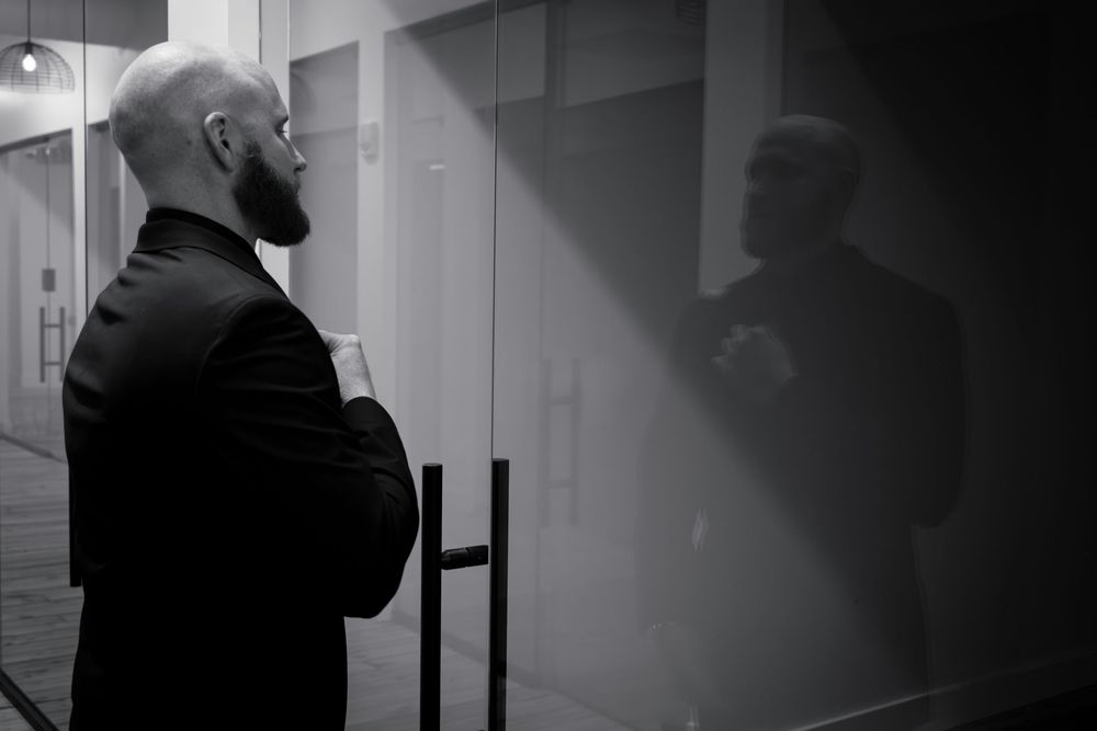 A person in a dark jacket stands in profile against a stark hallway with dramatic shadows and reflections.