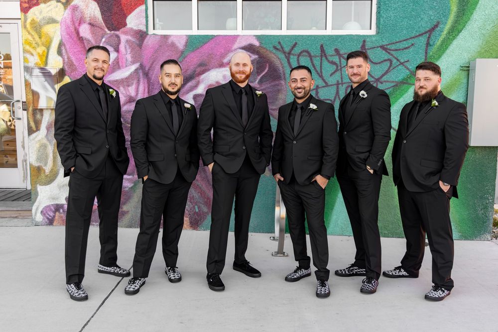 A row of groomsmen in black suits sit on a ledge showing off their colorful socks in a playful wedding photo.