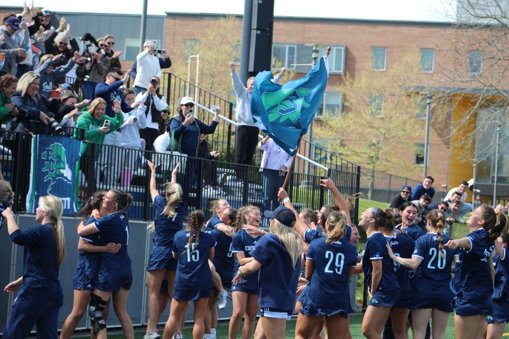 Team in navy uniforms celebrates with cheering fans waving flags in stadium stands.