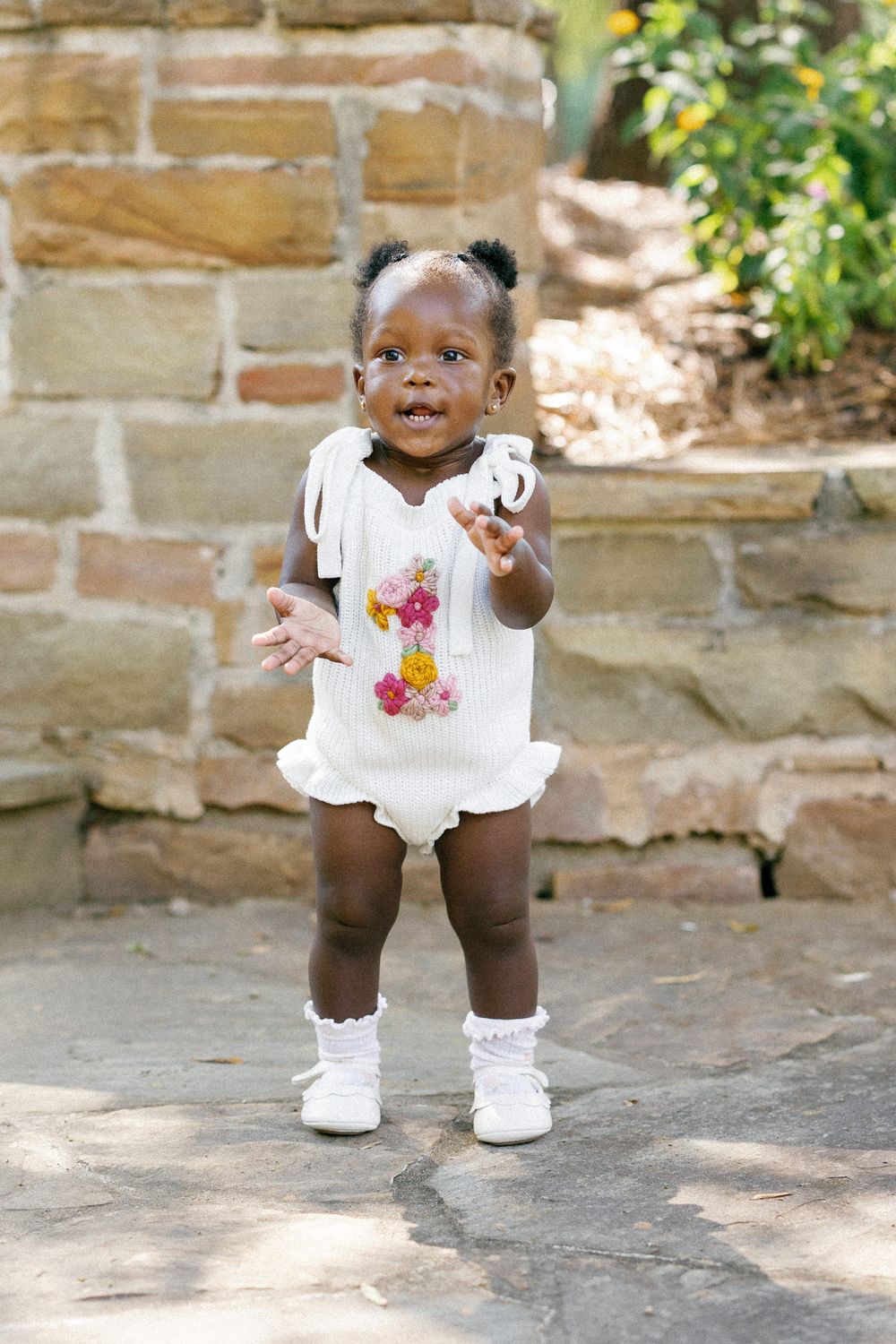A baby wearing a white floral dress and white shoes stands on stone pavement in a series of candid poses.