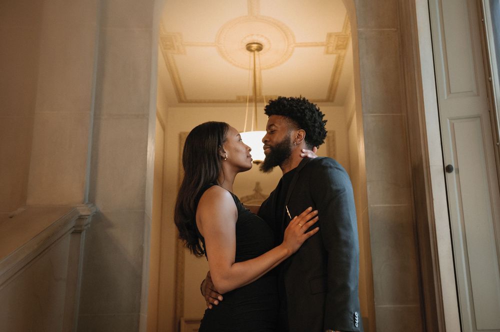 A romantic couple shares an intimate kiss in front of a white wall with elegant pendant lighting.
