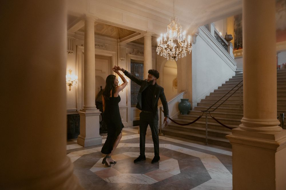 Two figures dancing elegantly in an ornate ballroom with a grand staircase and crystal chandelier.