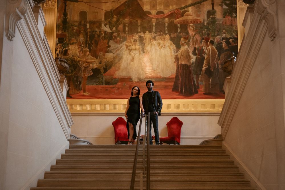 Two people stand in a grand museum hallway with large historical paintings on display.