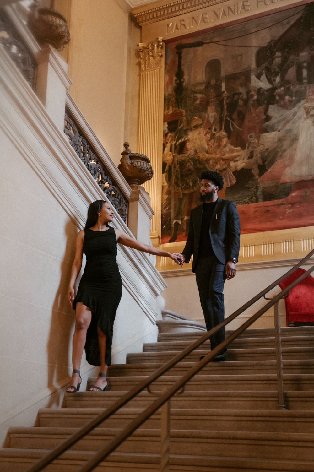 Two people descend a grand staircase in a historic building with classical artwork on the walls.
