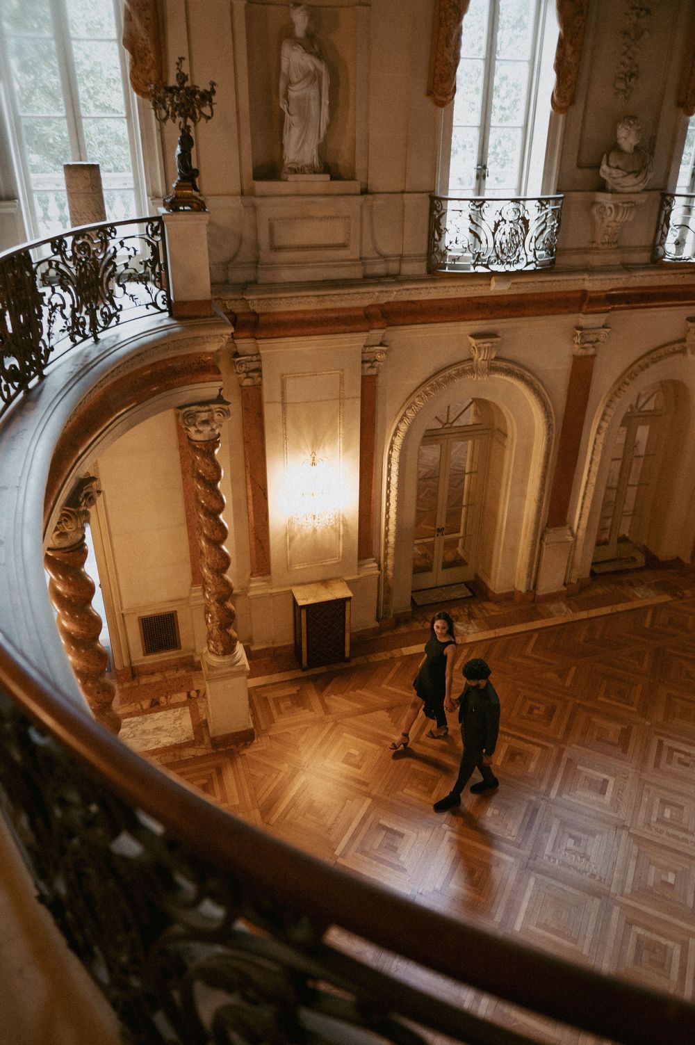 Couple standing in an elegant historic mansion with ornate architecture, spiral staircase, and classical statues.