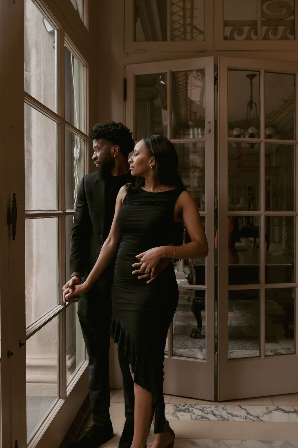 Dancers in black outfits perform intimate movements near large windows in a studio space.