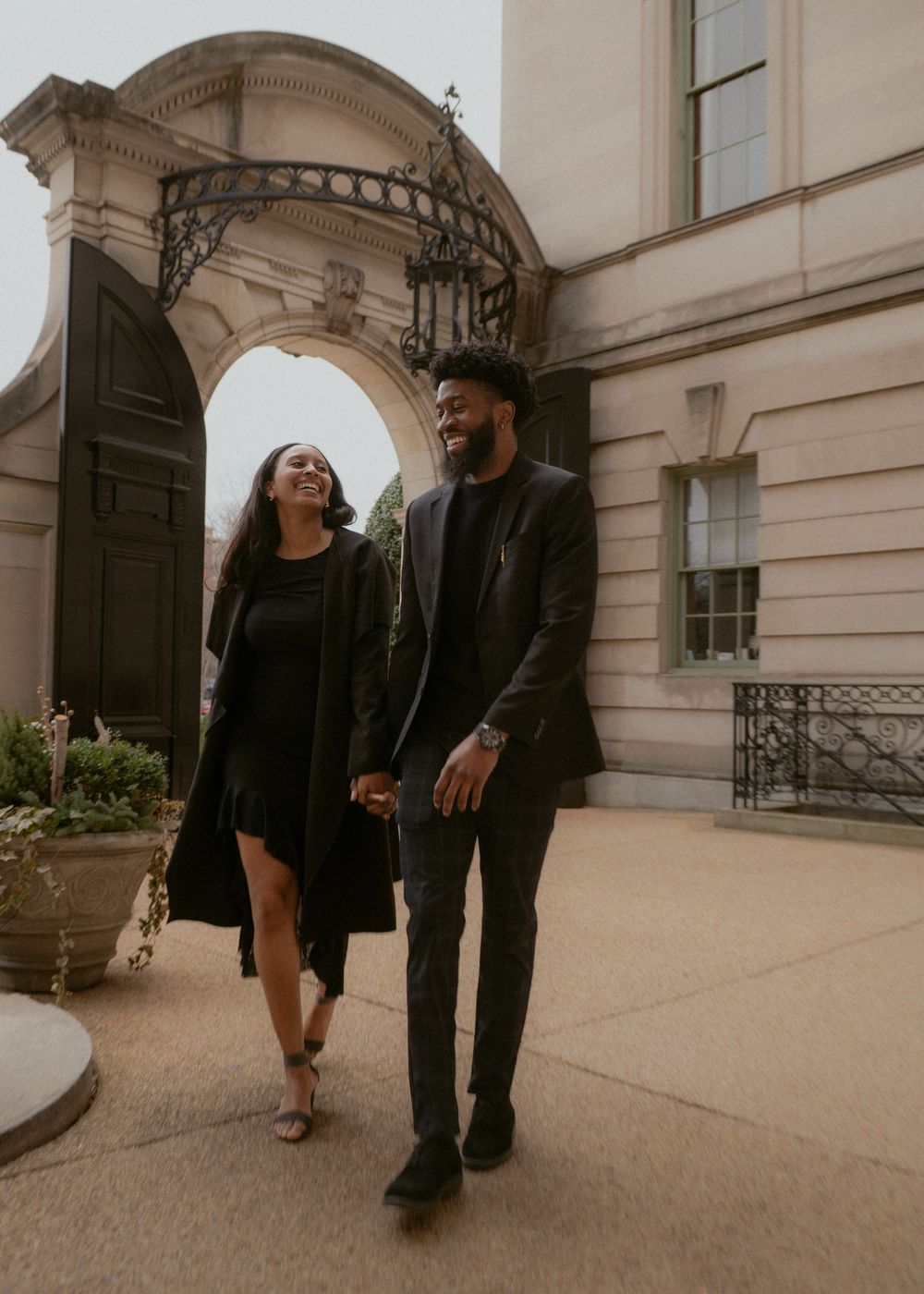 A couple walking and posing together on a beige stone terrace outside a classical building.
