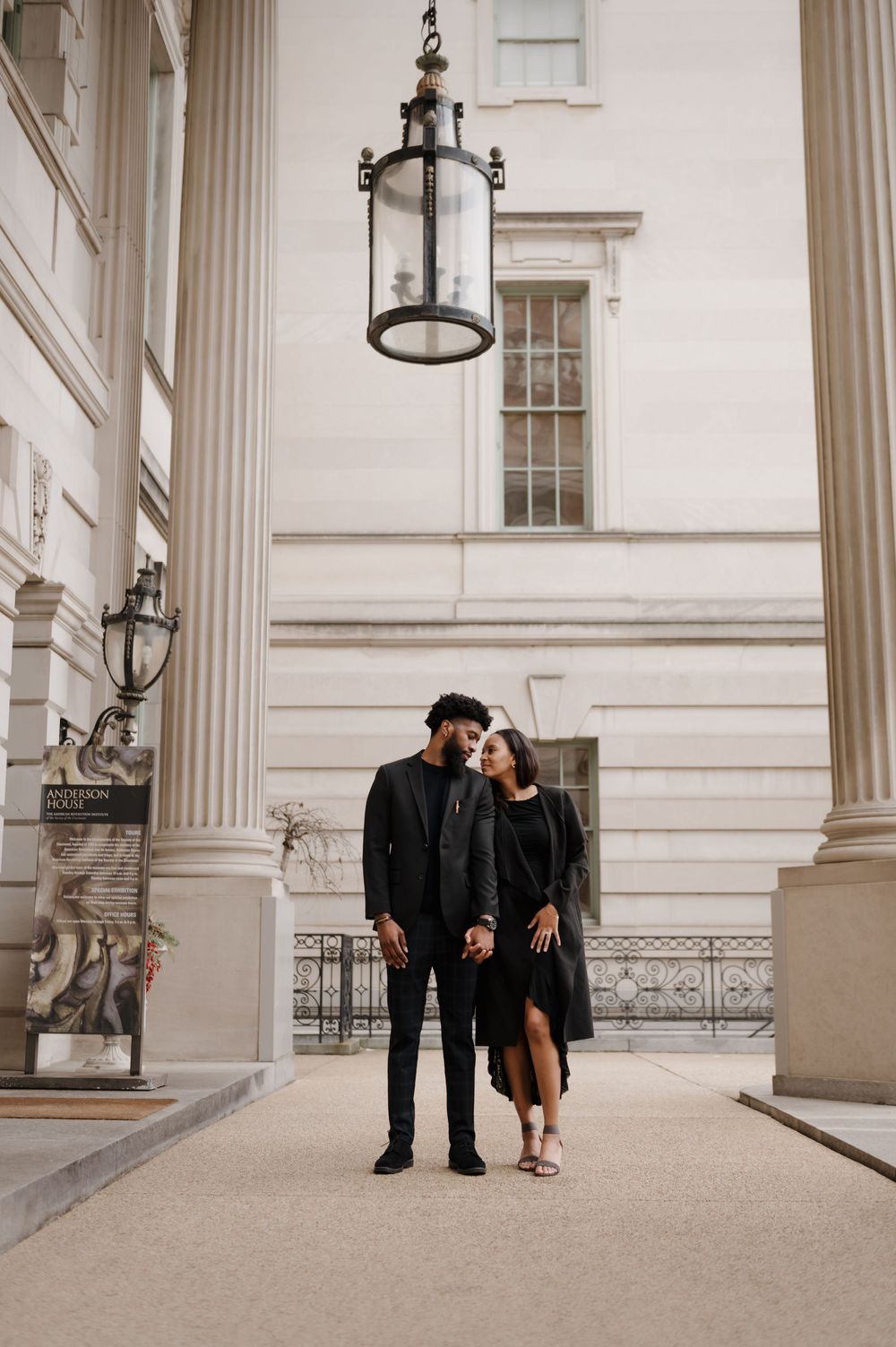 A couple stands beneath classical columns and hanging lanterns in formal attire.