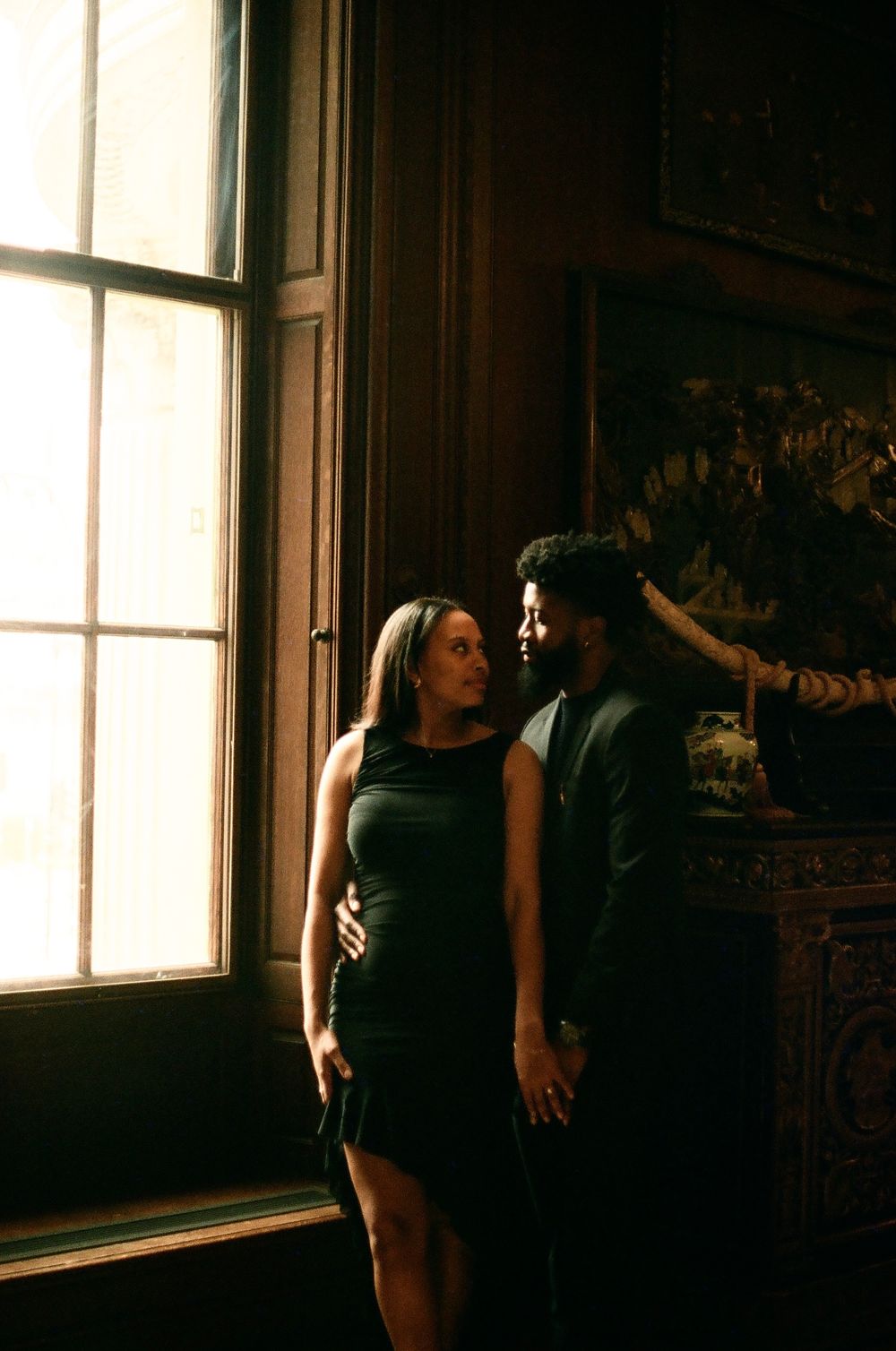 Figures pose in dramatic lighting near a tall window in a historic interior.