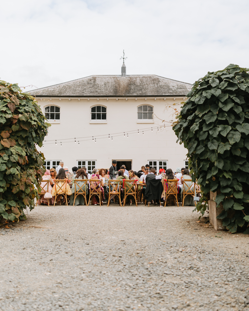 Long outdoor reception tables are set up at a rustic venue with climbing vines and gravel ground.