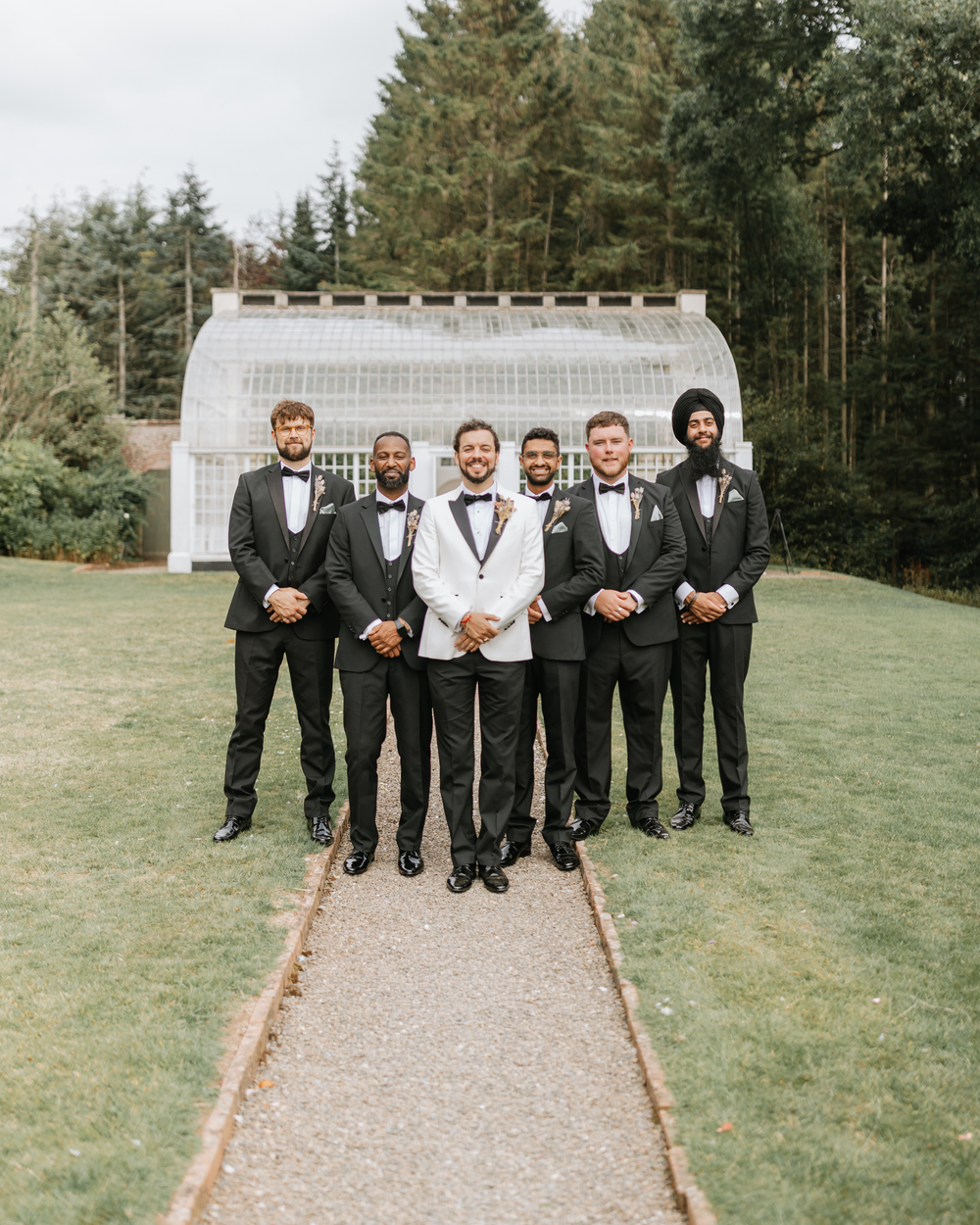 A group of groomsmen in black tuxedos and bowties pose together on a path surrounded by evergreen trees.