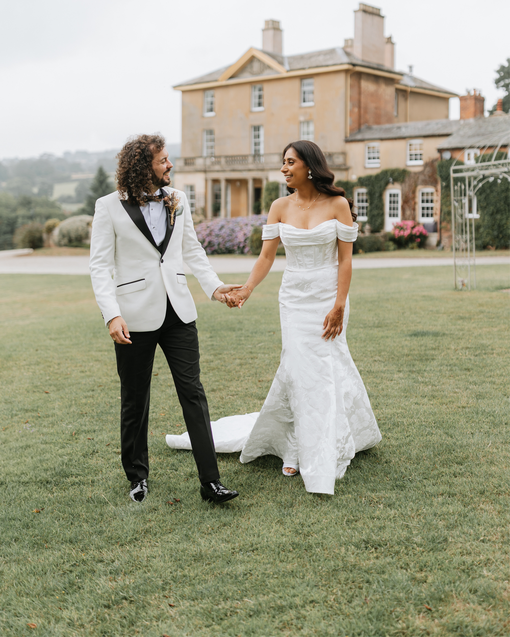 A couple in wedding attire dances playfully on a grassy lawn with a historic mansion in the background.