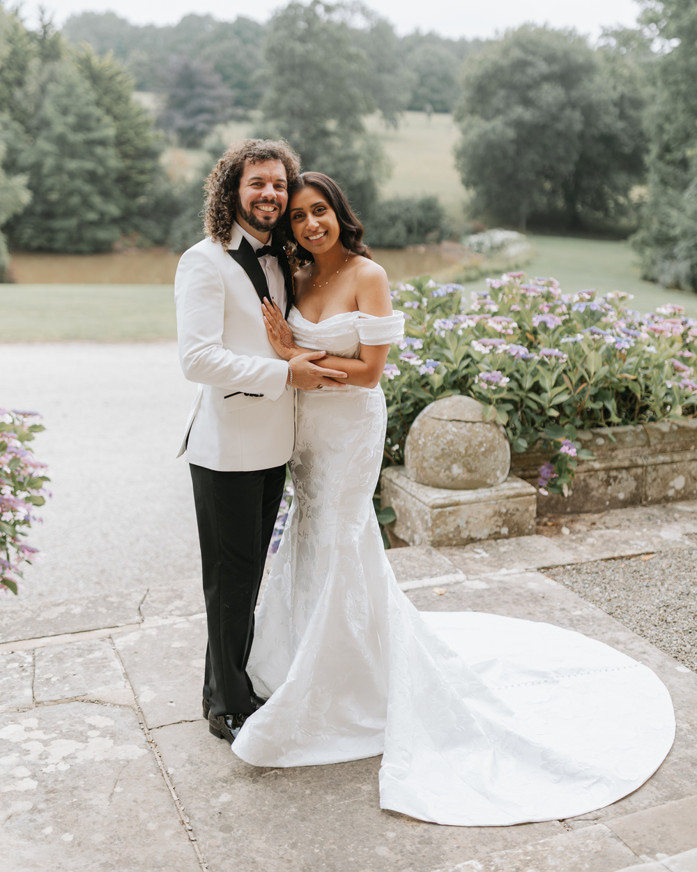 A couple in formal wedding attire dances together on a stone pathway surrounded by lush greenery and gardens.