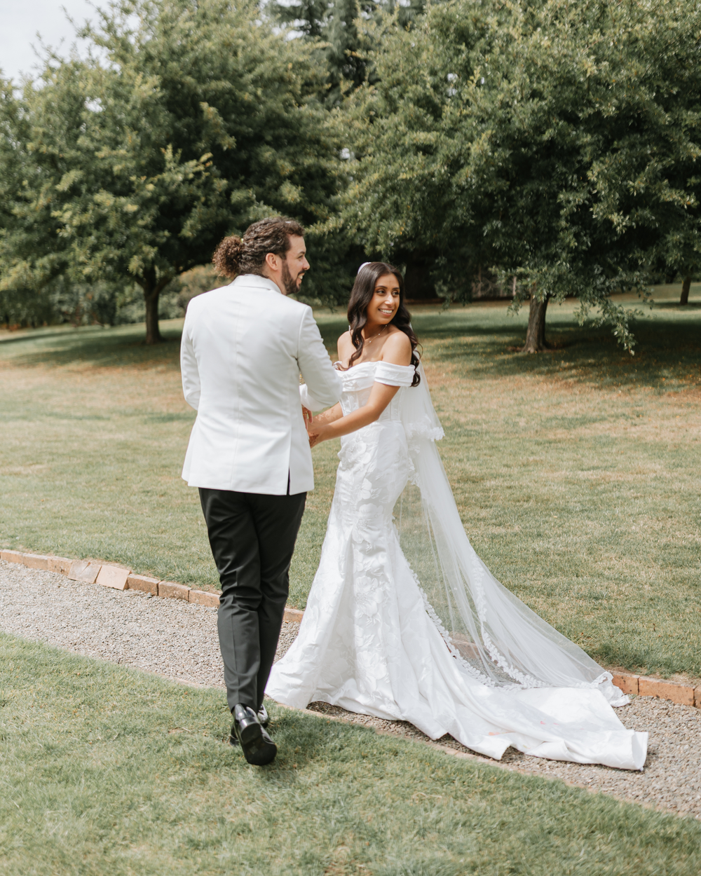 A couple in white and black formal wear twirls and dances together on a garden path surrounded by trees.
