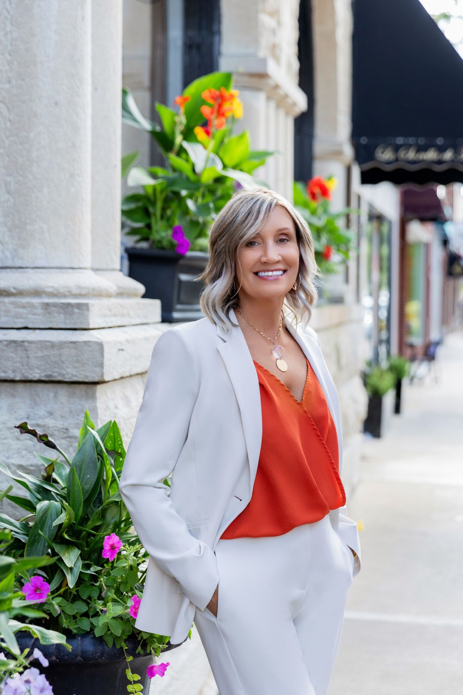 Professional woman in white suit and orange blouse smiles while standing by floral planters on city sidewalk.