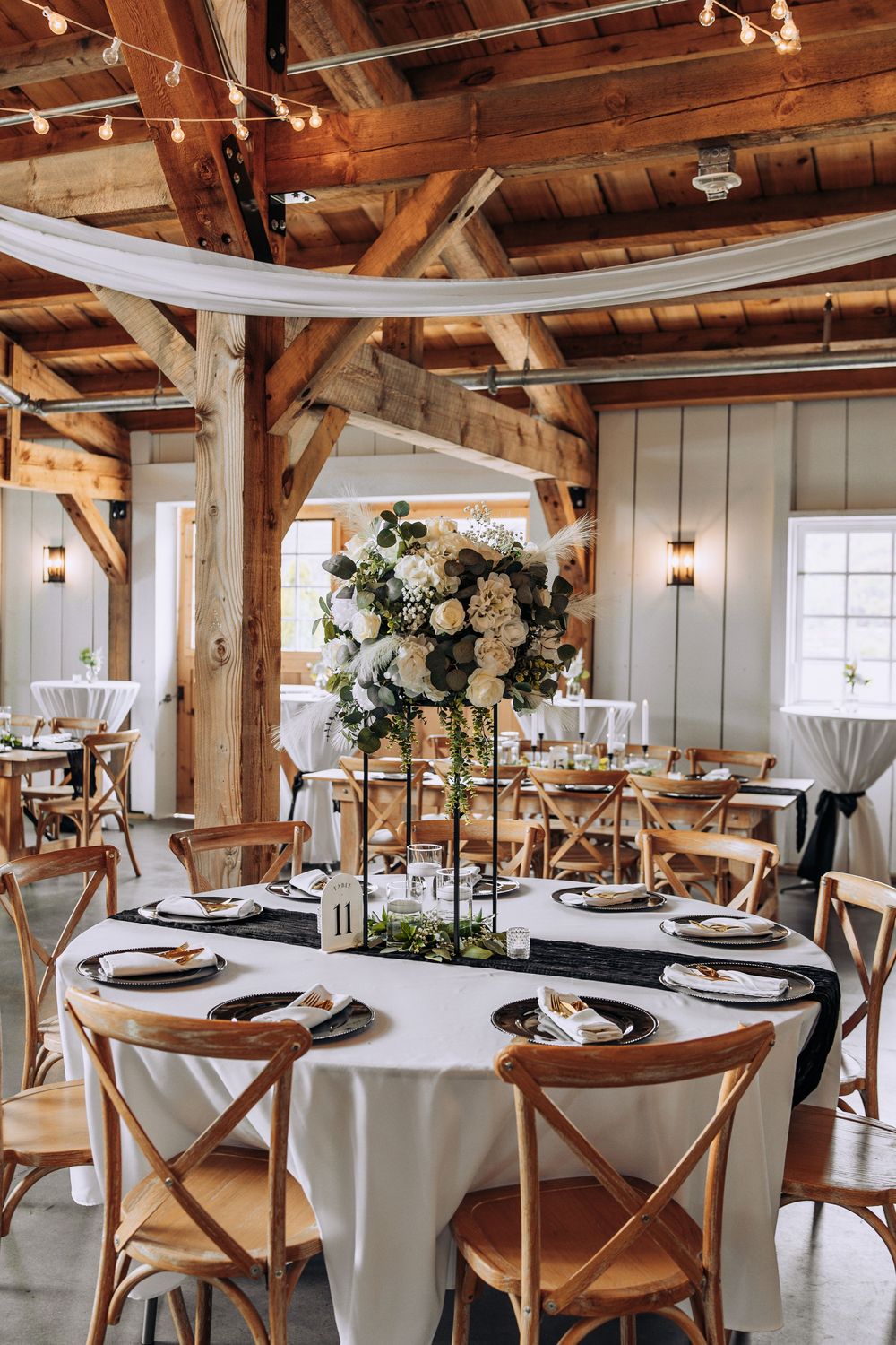 Elegant barn wedding reception table with tall white floral arrangement and cross-back chairs.