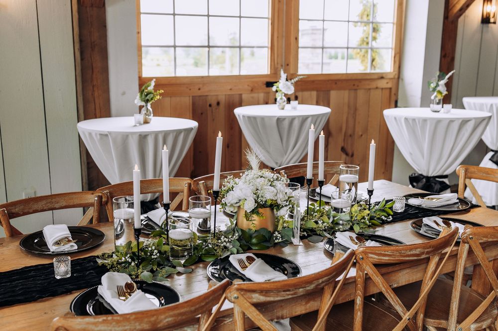 Elegant wedding reception table with white candles, greenery runner, black chargers, and floral centerpieces on a rustic wooden table.