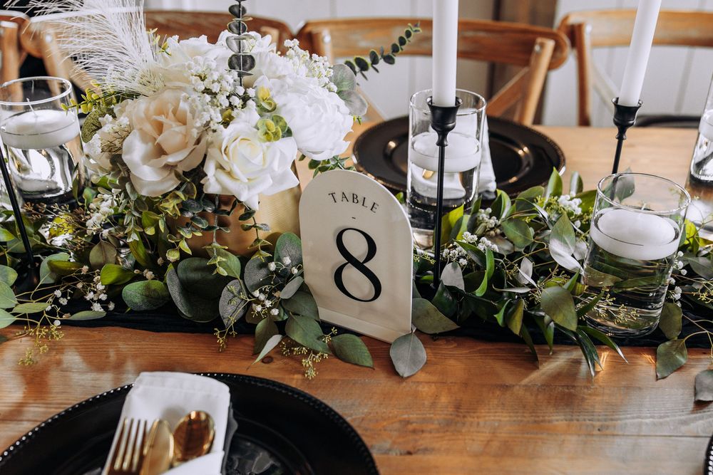 Wedding table centerpiece with white roses, greenery, black taper candles, and a Table 8 number card on a wooden table.
