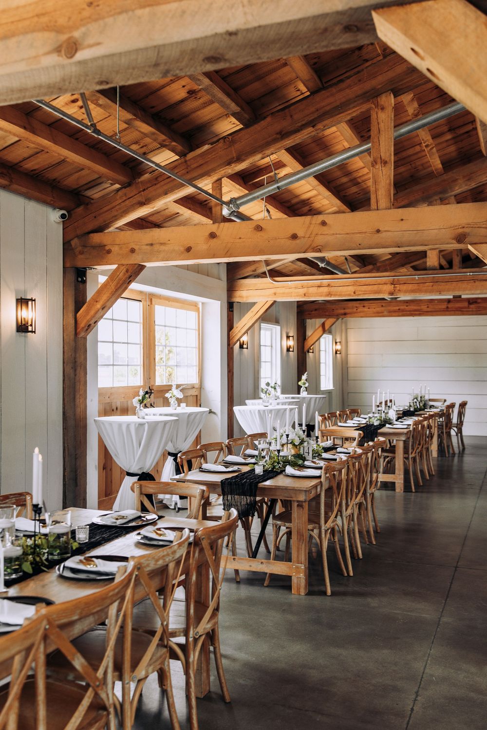 Rustic barn wedding venue with exposed wooden beams, long farm tables, cross-back chairs, and elegant white table settings.