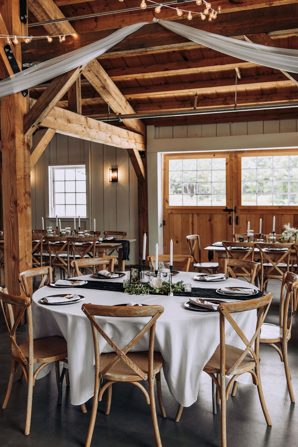 Rustic barn wedding reception with vaulted wood ceiling, round tables with white linens, cross-back chairs, and candle centerpieces.