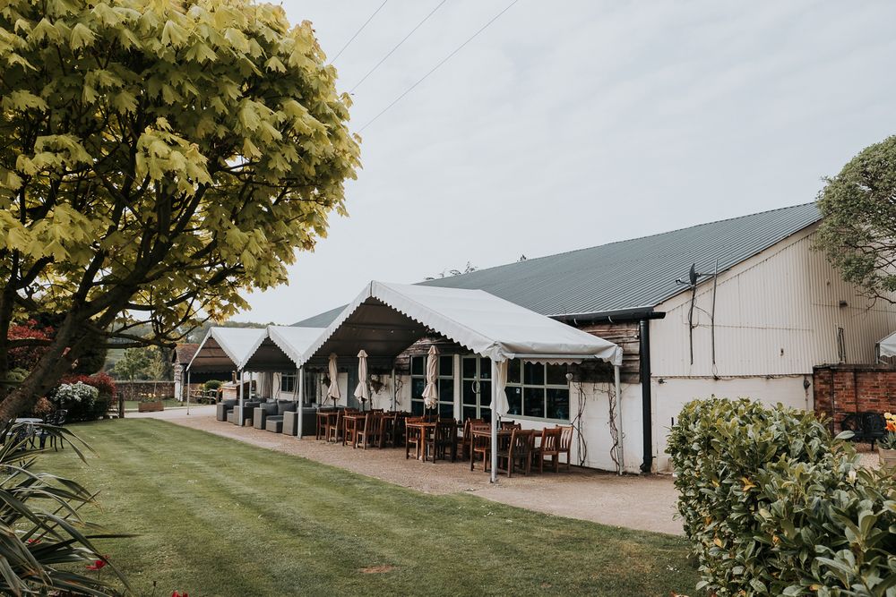A panoramic view of white wedding tents and barns set up on a grassy field with trees in the background.